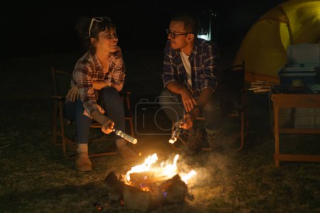 Téléchargez les photos : Asian couple roasting marshmallows together at a campfire where they set up a tent to camp by the lake at night. - en image libre de droit