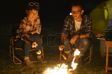 Téléchargez les photos : Asian couple is grilling corn and sausages at a campfire where they set up a tent to camp by the lake at night. - en image libre de droit
