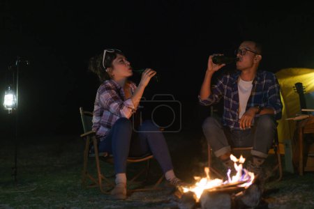 Téléchargez les photos : An Asian couple drinking beer with a campfire they set up to light the night while in the middle of a camping tent near a lake. - en image libre de droit