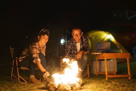 Téléchargez les photos : Asian couple grill corn on a fire they set up to light the night while in the middle of a tent camping near a lake. - en image libre de droit
