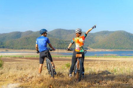 Téléchargez les photos : Asian cyclist couple looking at lake and mountain view and chatting while taking a break from the morning bike ride - en image libre de droit