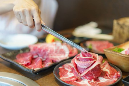 Die Hand der Frau setzt eine Zange ein, um Wagyu-Rindfleisch auf einen Teller zu legen, fertig zum Grillen über Holzkohle, was das kulinarische Erlebnis in einem japanischen Restaurant verbessert