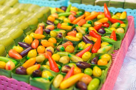 Téléchargez les photos : Un assortiment de fruits en forme de haricots mungs desserts thaïlandais vibrants, conçus pour ressembler à des fruits et légumes miniatures, exposés dans des récipients clairs sur un marché - en image libre de droit