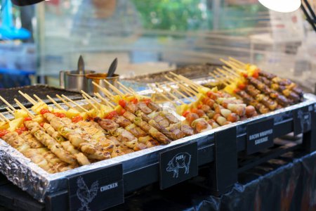Téléchargez les photos : Une gamme appétissante de brochettes grillées avec du poulet, du porc et des saucisses enveloppées de bacon, accompagnées de légumes, exposées dans un kiosque de nourriture de rue - en image libre de droit