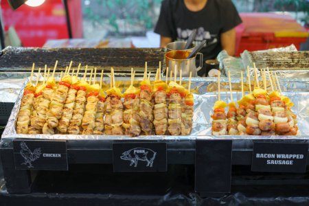 An appetizing array of grilled skewers featuring chicken, pork, and bacon-wrapped sausages, accompanied by vegetables, displayed at a street food stand