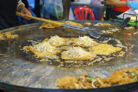 Thai street food chef preparing a traditional crispy oyster omelette, garnished with fresh bean sprouts on a large iron griddle