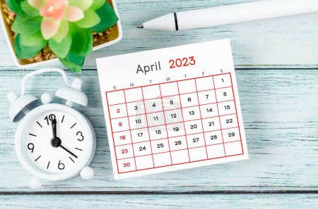 April 2023 Monthly calendar year and alarm clock with pen on blue wooden background.