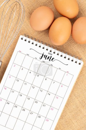 June 2024 Desk calendar and fresh eggs with egg whisk on burlap background.