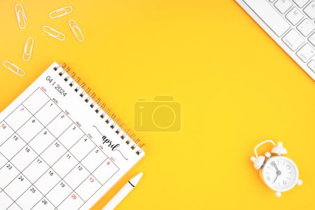 April 2024, Monthly desk calendar for 2024 year and alarm clock with paper clips on yellow color background. Position with copy space.