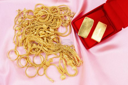 Many gold necklaces and gold bars in red box on pink velvet cloth background.