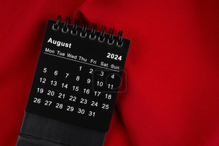 August 2024 Black spiral desktop calendar on red textile.