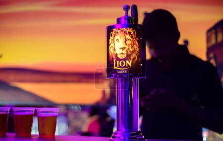 Foto de Weligama, Sri Lanka - 09 29 2022: Lion lager backlit logo on a liquor pub table and 3 beer cups. Silhouetted bartender in the background. - Imagen libre de derechos