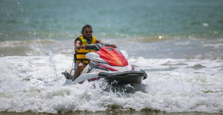 Foto de Weligama, Sri Lanka - 10 29 2022: Handsome Jetsky rider wearing sunglasses and a yellow jacket riding a jet ski on the beach. - Imagen libre de derechos
