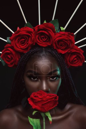 Closeup of a young black woman holding red flower is looking at camera.  Portrait of a beautiful young woman indoors in studio on a black background with flowers on her head, with attractive eyes.