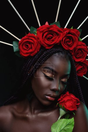 Photo for Young black woman has her eyes closed tilted her head holding a red flower.  Close-up of beautiful young black woman is with flowers on her head, in black studio background. - Royalty Free Image
