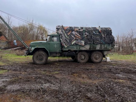 Foto de Gran camión militar cubierto con red de camuflaje contra el fondo de una antigua valla y tierra sucia de la lluvia. Guerra en Ucrania - Imagen libre de derechos