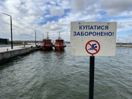 Foto de Signo de prohibición en el río. Traducción: "Nadar está prohibido". En el fondo (fuera de foco) hay dos botes de bomberos y un río de verano - Imagen libre de derechos