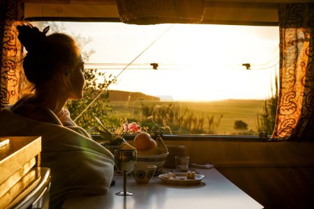 the girl sits looking at the sunset through the window in the camper. Caravan