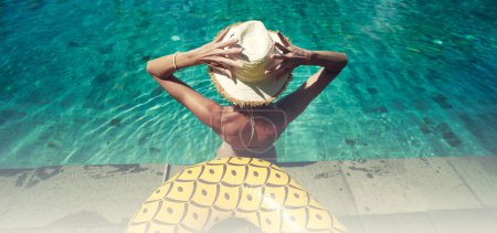 Photo for Back view of young woman wearing summer hat relaxing in big swimming pool with blue water on a sunny day. Copy space. Luxury hotel - Royalty Free Image