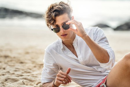 Foto de Retrato de un joven italiano guapo relajándose en la playa, usando camisa blanca y gafas de sol de moda. Hora de verano. - Imagen libre de derechos