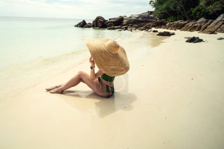 Foto de Photo fo a woman enjoying sunbath at tropical beach, sitting on the sand, wearing big summer hat. Tanned girl relaxing on vacation. Tourist. Island. - Imagen libre de derechos