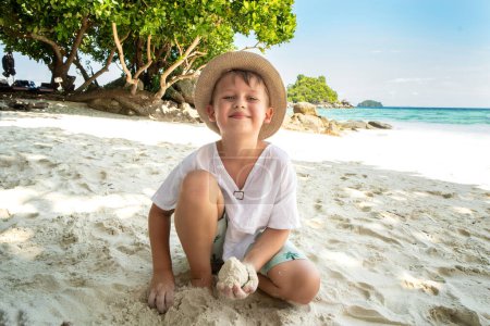 Téléchargez les photos : Little happy boy in summer hat playing with sand on the tropical beach, looking at the camnera with big smile on his face. Beautiful landscape. Blue sky and sea water background. Kid on vacation. - en image libre de droit