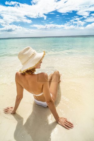 Photo for Woman relaxing on the tropical beach, sitting on the sand, sunbathing. Travel concept. Girl in white bikini and hat. Dream island. Summer vibes - Royalty Free Image