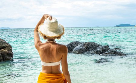 Téléchargez les photos : Traveler girl relaxing on a tropical beach, in nature. Summer vacations. Back view of beautiful woman in swimsuit and hat spending leisure time, enjoying sunny day. Wanderlust. - en image libre de droit