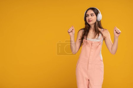 Téléchargez les photos : Cheerful young woman wearing headphones, listening music or podcast, yellow studio background. A lot of copy space. Girl dancing, relaxing. Real people. - en image libre de droit