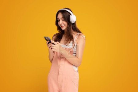 Téléchargez les photos : Smiling female student listening to music in bluetooth headphones and using smartphone. Studio shot, yellow background, Happy young woman. Online app in mobile phone. Copy space. - en image libre de droit