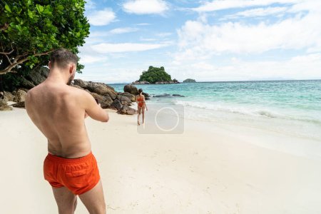 Foto de Man using smartphone camera, taking photo to a beautiful woman at the tropical beach. Vacation time. Summer travel. Tourism. Tourists - Imagen libre de derechos