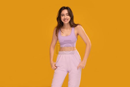 Photo for Studio photo of a happy, relaxed, young brunette woman in sporty clothes posing over yellow background with big smile, looking at the camera. Female student. A lot of copy space - Royalty Free Image