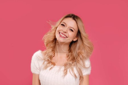 Foto de Young adult woman with long blond wavy hair smiles happily while looking at the camera. Beauty portrait. Pastel pink studio background. - Imagen libre de derechos