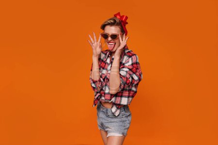 Téléchargez les photos : Photo of crazy pinup girl in checkered shirt and short jeans smiling and showing tongue out. Retro vintage 50s style, studio shot isolated on orange background. A lot of copy space - en image libre de droit