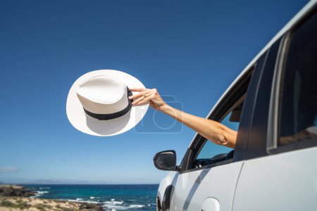 Téléchargez les photos : Femmes main tenant chapeau blanc à l'extérieur de la fenêtre de voiture ouverte. Concept de voyage. Rêve. La soif d'errance. Tourisme. Espace de copie. - en image libre de droit