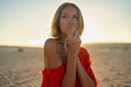 Foto de Retrato de una romántica mujer rubia con un vestido rojo posando en el desierto de arena a la luz dorada del atardecer. Chica con anillos de moda en las manos. - Imagen libre de derechos