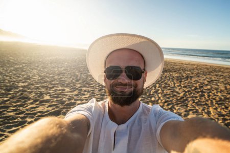Foto de Feliz, sonriente hombre de vacaciones en la playa, tomando selfie. Chico guapo en sombrero de verano blanco y gafas de sol negras. - Imagen libre de derechos