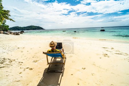 Téléchargez les photos : Homme au chapeau d'été, homme d'affaires, nomade numérique travaillant avec un ordinateur portable sur la plage tropicale. Style de vie freelance. Les tropiques. Ambiances estivales - en image libre de droit