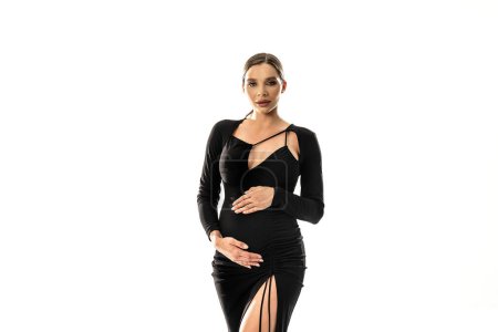 Téléchargez les photos : Concept de grossesse. Femme posant en fond de studio blanc portant une robe noire, tenant son ventre enceinte, regardant la caméra. Beaucoup d'espace de copie. - en image libre de droit