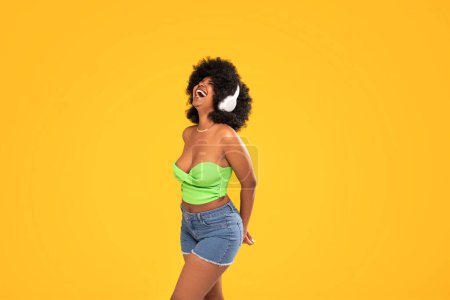 Téléchargez les photos : Adorable femme afro-américaine avec des écouteurs blancs riant à haute voix, dansant, fond studio vibrant jaune. De vraies émotions. Beaucoup d'espace de copie. - en image libre de droit