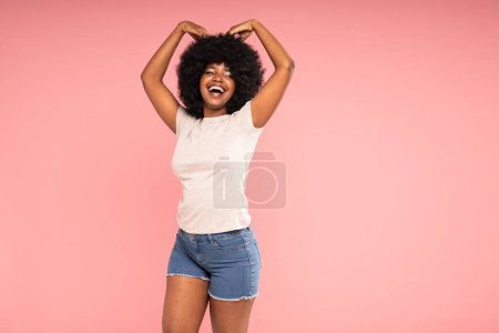 Photo for Charismatic happy natural african american woman with afro hairstyle dancing and laughing out loud, posing on the pink pastel studio background. Real people emotions. A lot of copy space. - Royalty Free Image