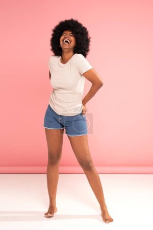Téléchargez les photos : Femme optimiste afro-américaine qui s'amuse, sourit, rit joyeusement. Fille avec une coiffure afro portant un t-shirt décontracté et un jean court, posant sur un fond rose pastel. - en image libre de droit