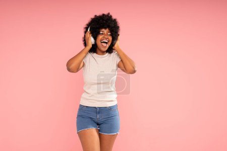 Téléchargez les photos : Joyeux jeune femme avec afro coiffure danse, écouter de la musique dans les écouteurs, s'amuser seul. Fille heureuse posant sur fond de studio pastel rose. - en image libre de droit