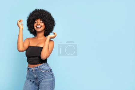 Téléchargez les photos : Portrait de jeune femme afro-américaine joyeuse modèle en haut noir et jeans, avec coiffure afro, souriant à la caméra. De vraies émotions. Beaucoup d'espace pour copier. Bleu pastel fond studio. - en image libre de droit