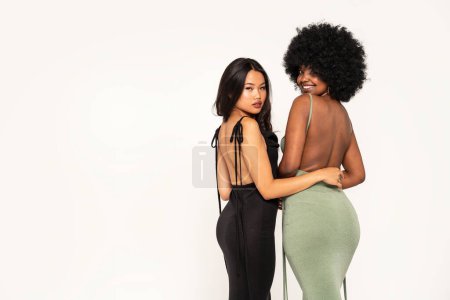 Photo for Fashionable two women posing in elegant party dresses, having fun together, looking at the camera and smiling. Back view. Studio Background. Real people lifestyle. Body positive and conscious. A lot of copy space - Royalty Free Image