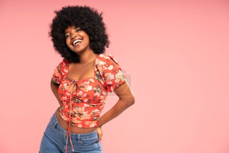 Photo for Charming girl looking and smiling to the camera, posing in jeans and fashionable top. Attractive laughing african woman. Pink pastel studio background. Copy space. - Royalty Free Image