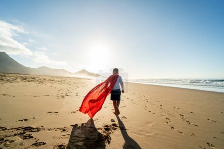 Photo for Superhero child walking on the sandy, sunset beach, wearing red cape. Back view. Full length. Summer vacation concept. A lot of copy space. Kids power. Happiness. - Royalty Free Image