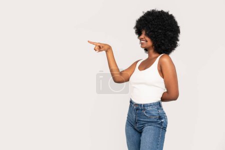 Téléchargez les photos : Heureuse fille afro-américaine avec une coiffure afro et un grand sourire pointant sur le fond vide du studio. Jeune femme en haut blanc et en jeans. Concept de vente - en image libre de droit