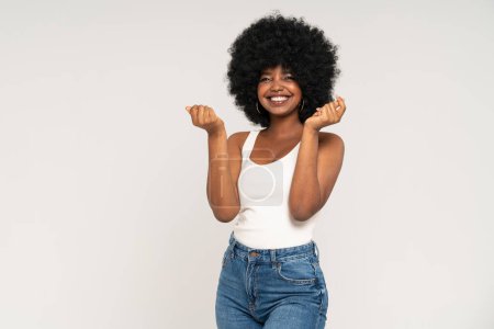 Téléchargez les photos : Heureuse fille afro-américaine avec une coiffure afro et un grand sourire posant sur un fond de studio gris clair. Jeune femme en haut blanc et en jeans. Concept de vente - en image libre de droit