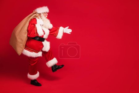 Photo for Funny old Santa Claus on a red studio background. Merry Christmas and Happy New Year! Copy space. Xmas sale, discount concept. Full length photo. - Royalty Free Image